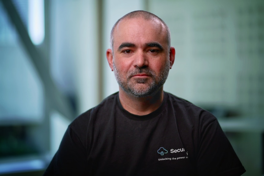 A middle aged man with a buzzed head and grey stubble wearing a black t-shirt looks at the camera with a neutral expression.