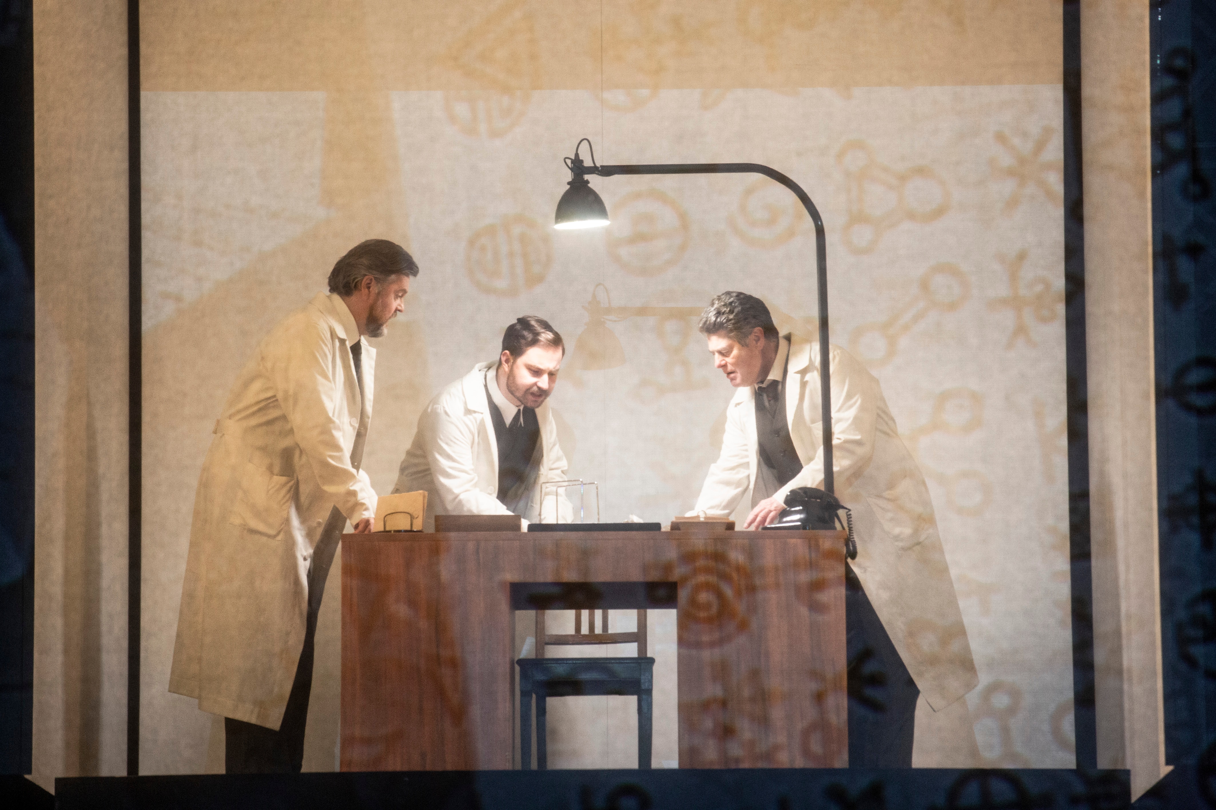 Three men dressed in lab coats gathered around a desk looking at something.
