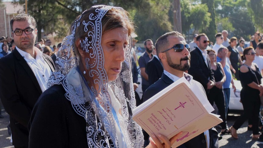 A woman stands in a crowd and reads from a booklet.