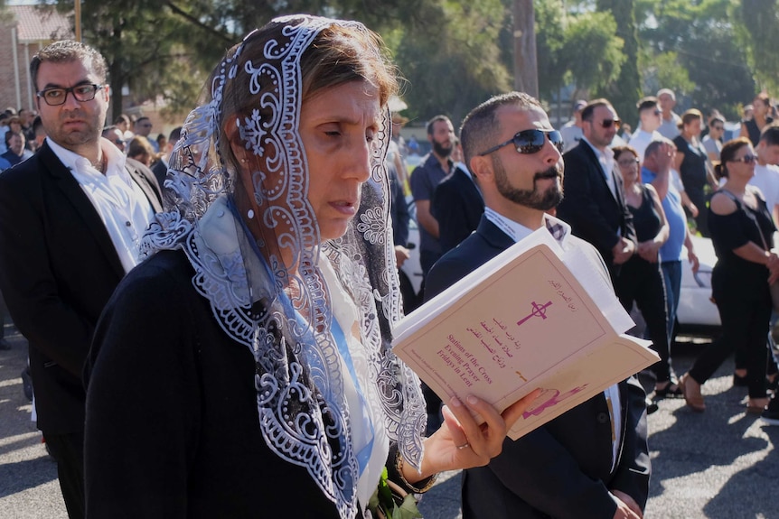 A woman stands in a crowd and reads from a booklet.