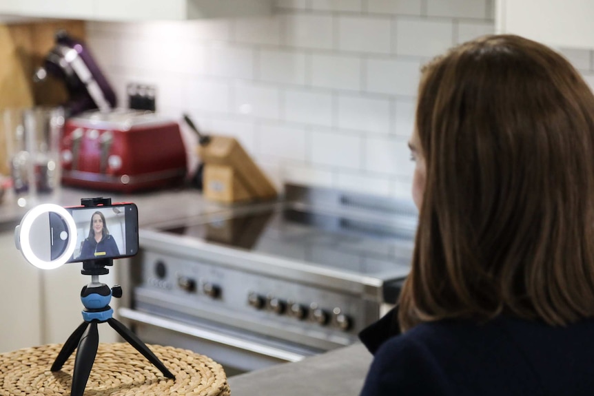 Henderson in kitchen looking at iphone on tripod with her image on it.