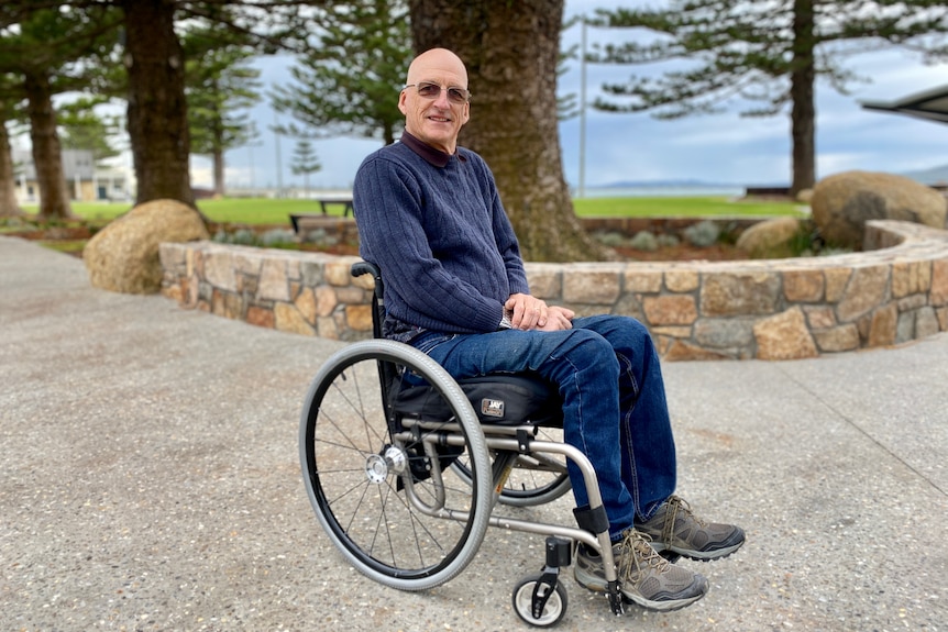 Greg Cross sits in his wheelchair on a footpath with grass and the beach in the background.