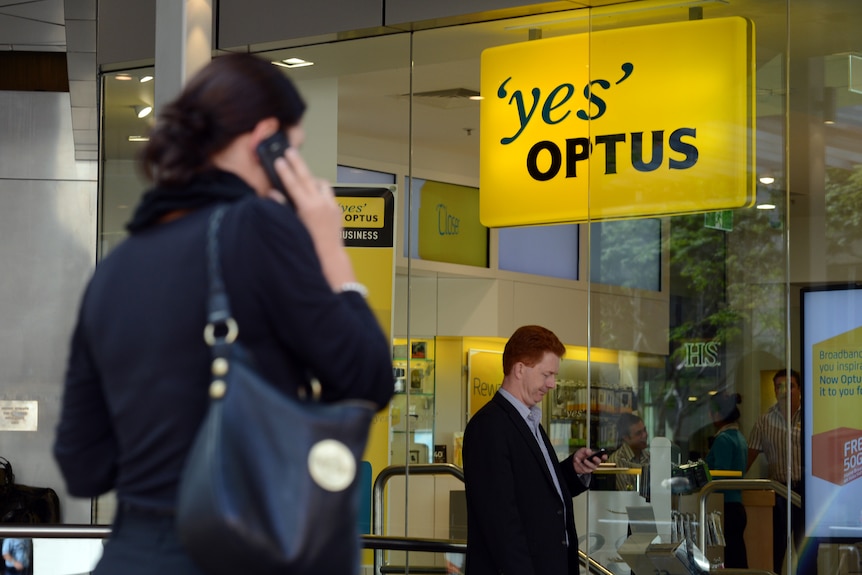 A woman uses her phone outside an Optus store while a man looks at his phone.