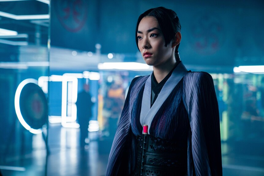An Asian woman in a galactic blue robe stands in a room with reflective panels and looks disapprovingly at something off camera.