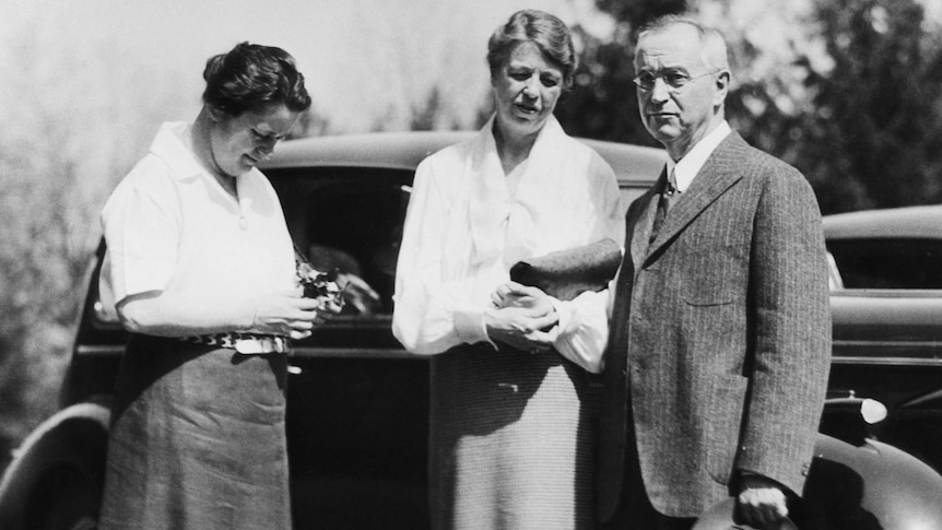 Three figures, two women and a man, stand in 1930s garb in front of a black car.