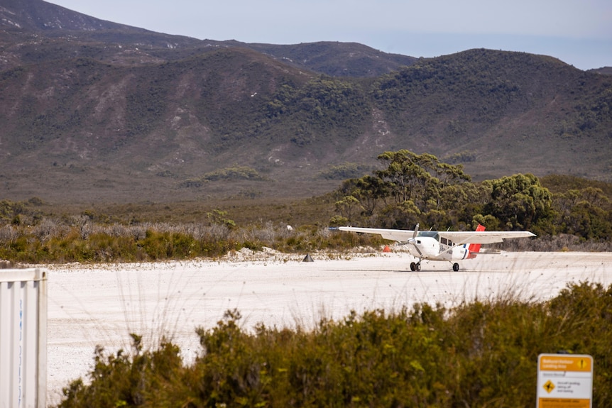 A small airplane with a propeller at the front lands on a white gravel airstrip with mountains in the background