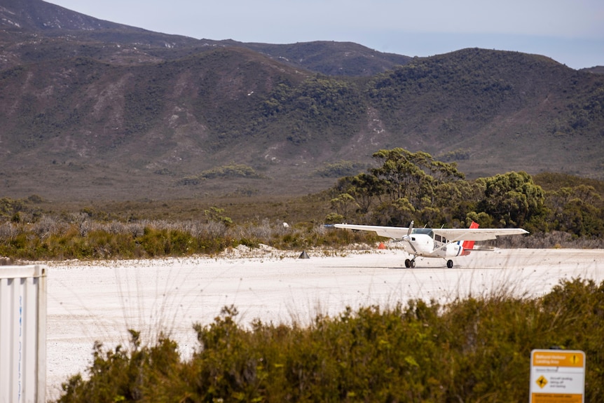 Un pequeño avión con una hélice en la parte delantera aterriza en una pista de aterrizaje de grava blanca con montañas al fondo