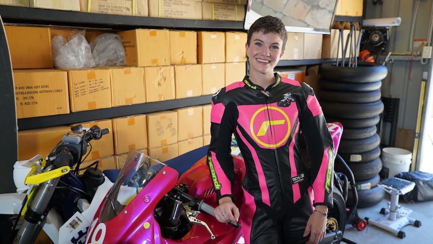 woman wearing motorbike leathers with pink accents leaning on a pink motorbike in a workshop 
