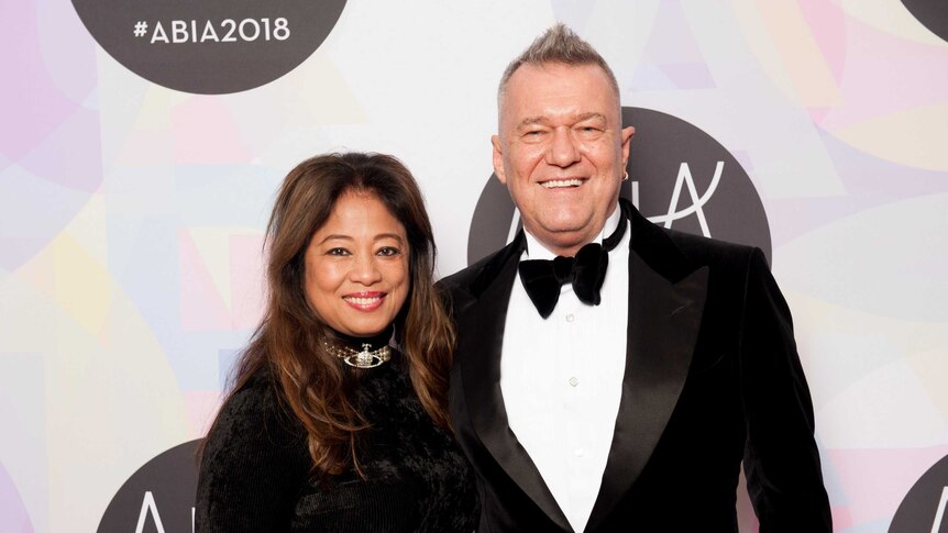 Barnes and Mahoney stand, smiling and wearing formalwear, arm in arm in front of a pastel coloured media wall.