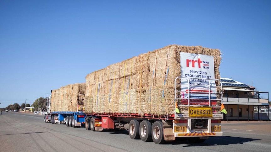 A truck full of hay travels through a small country town, with a sign on the back saying "providing drought relief to farmers...