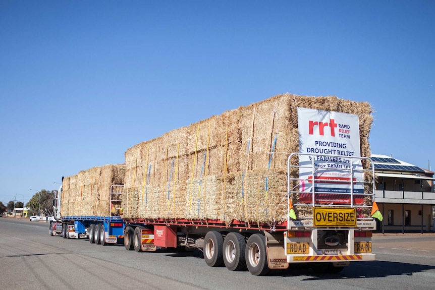 A truck full of hay travels through a small country town, with a sign on the back saying "providing drought relief to farmers...