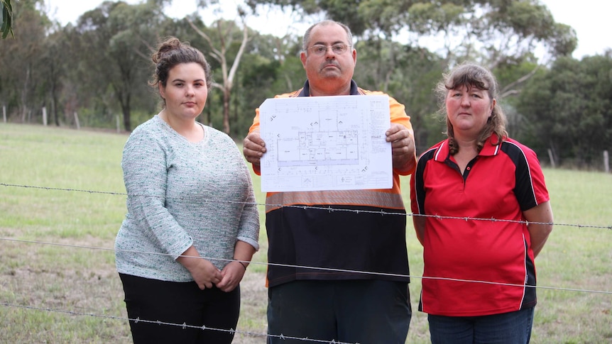 Emily, Kurt and Ann Pritchett standing behind a barbed wire fence, holding a house floorplan.