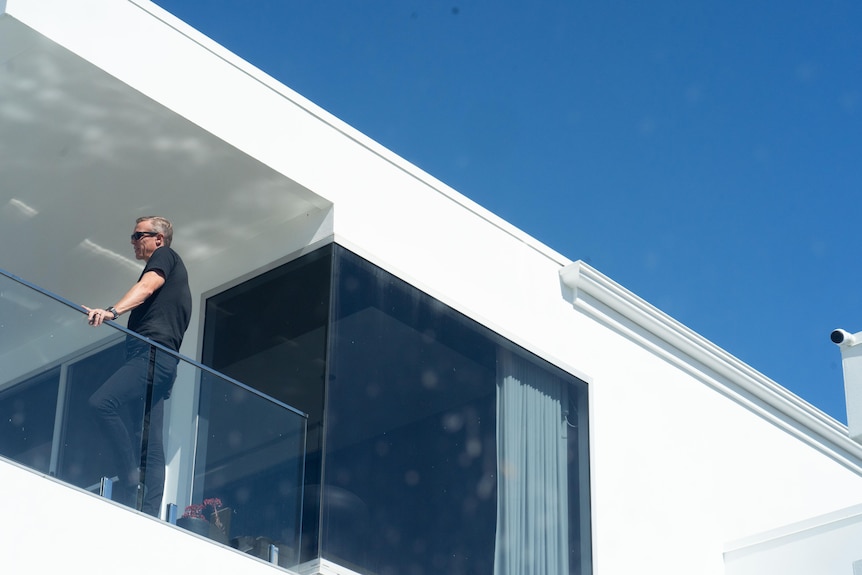 A man in a black shirt leans on the glass railing of a second level balcony