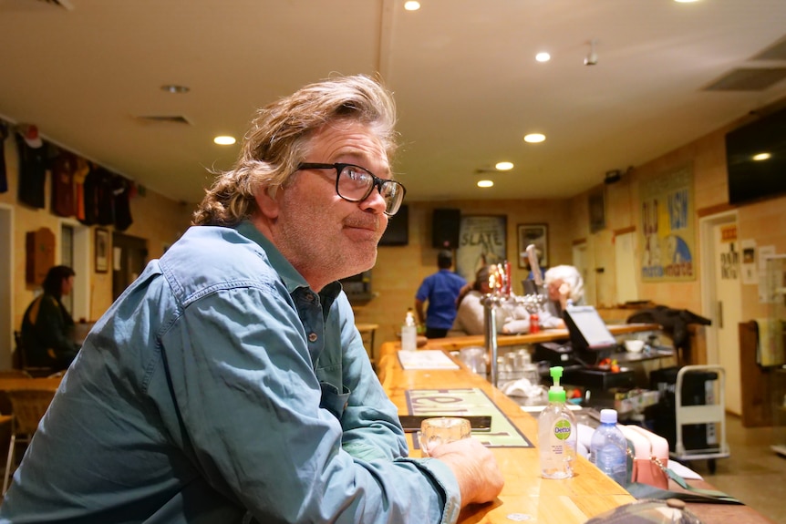 A man with glasses leans on the bar of a pub