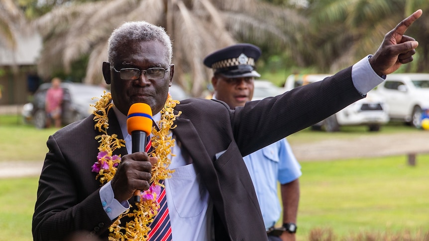 A man wearing a suit and a flower garland around his neck holds a microphone and points upwards.