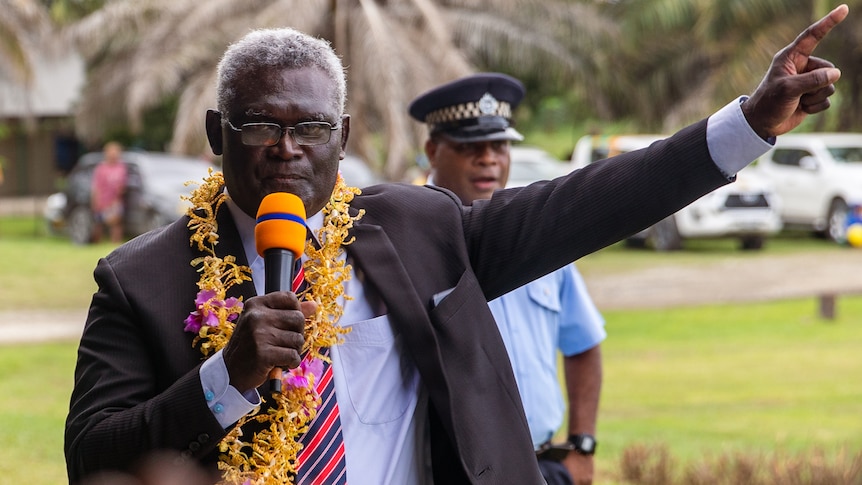 A man wearing a suit and a flower garland around his neck holds a microphone and points upwards.