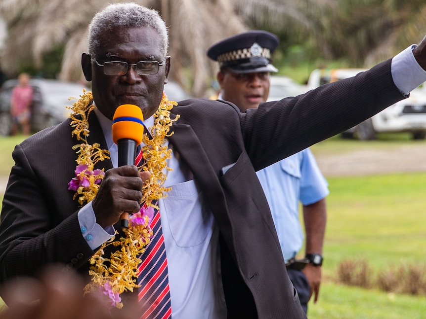 A man wearing a suit and a flower garland around his neck holds a microphone and points upwards.