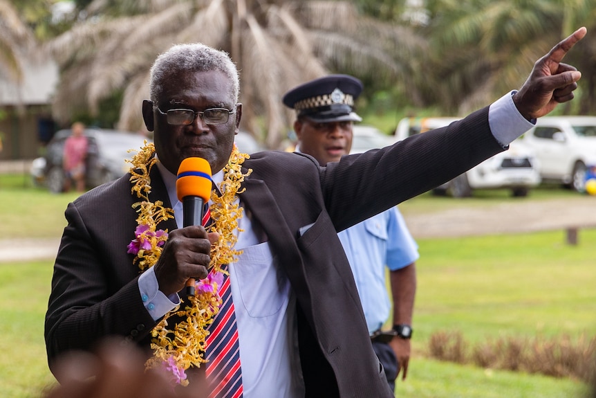 A man wearing a suit and a flower garland around his neck holds a microphone and points upwards.