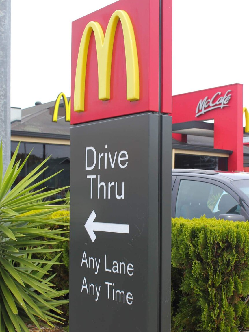A sign with a yellow M on a red background, below the sign reads 'Drive thru Any Lane Any time' a black car is in the background