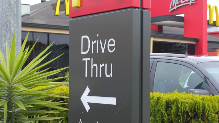 A sign with a yellow M on a red background, below the sign reads 'Drive thru Any Lane Any time' a black car is in the background