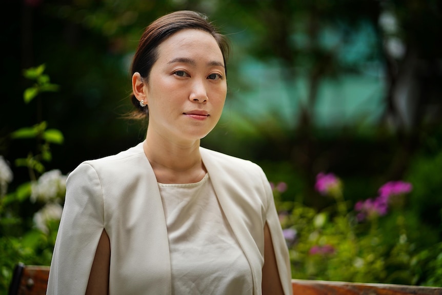 A Korean woman in a beige cape-style blazer standing solemnly in a garden 