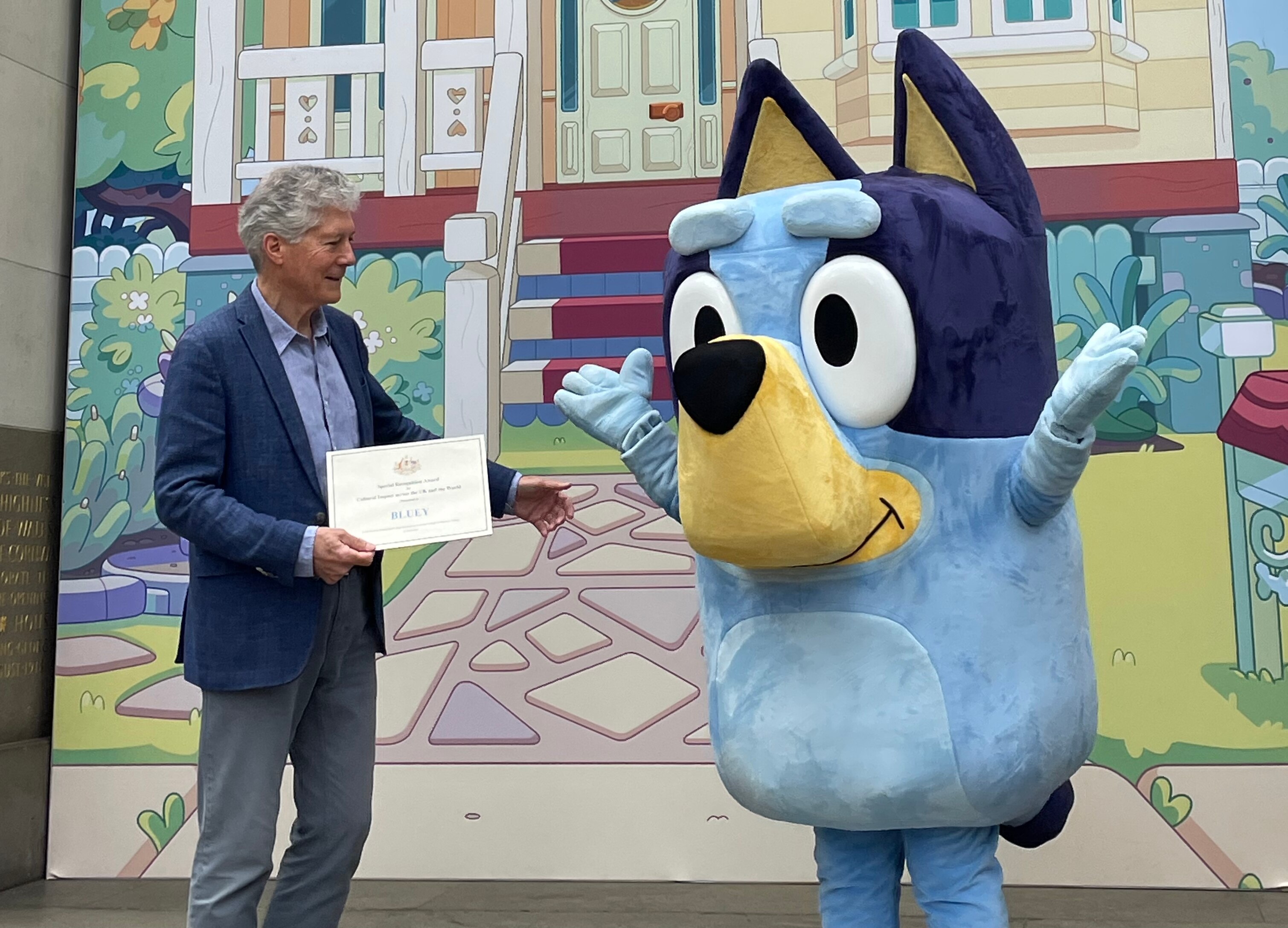 A man hands over a certificate to a lifesize mascot of Bluey, a blue dog.
