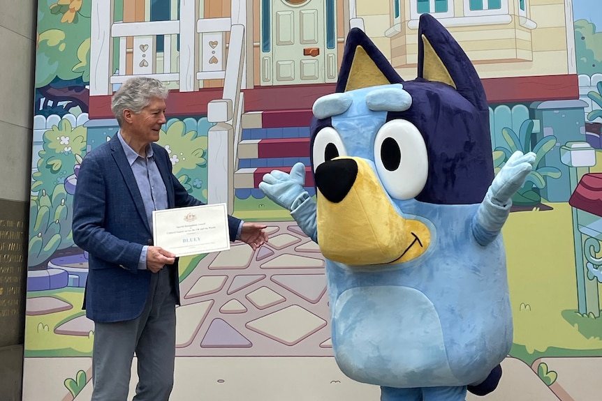 A man hands over a certificate to a lifesize mascot of Bluey, a blue dog.