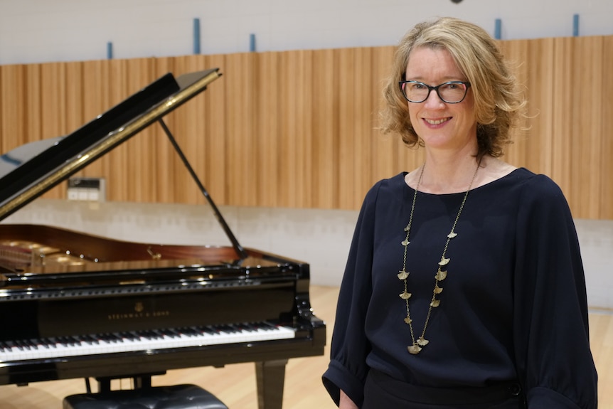 a woman in a dark top and long necklace is standing to the right of a shiny black grand piano