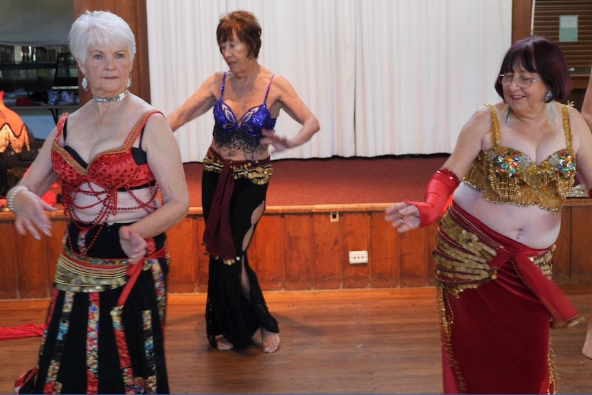 Three women in their late 60's and 70s with bellydance costumes on in dance poses.