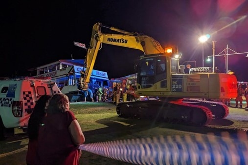 An excavator raising a boat that's taking on water late at night, with people gathered around watching 