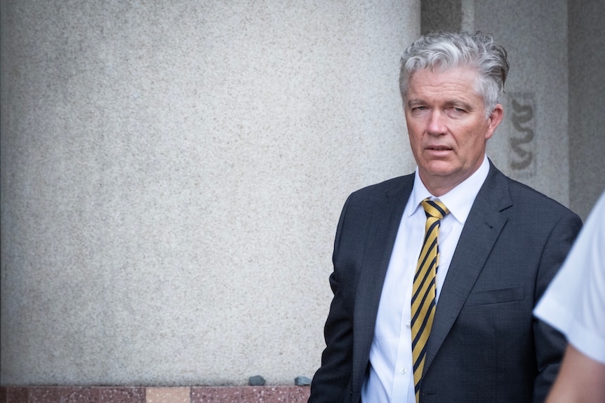 A grey-haired, middle-aged man in a dark suit walks away from a court building.