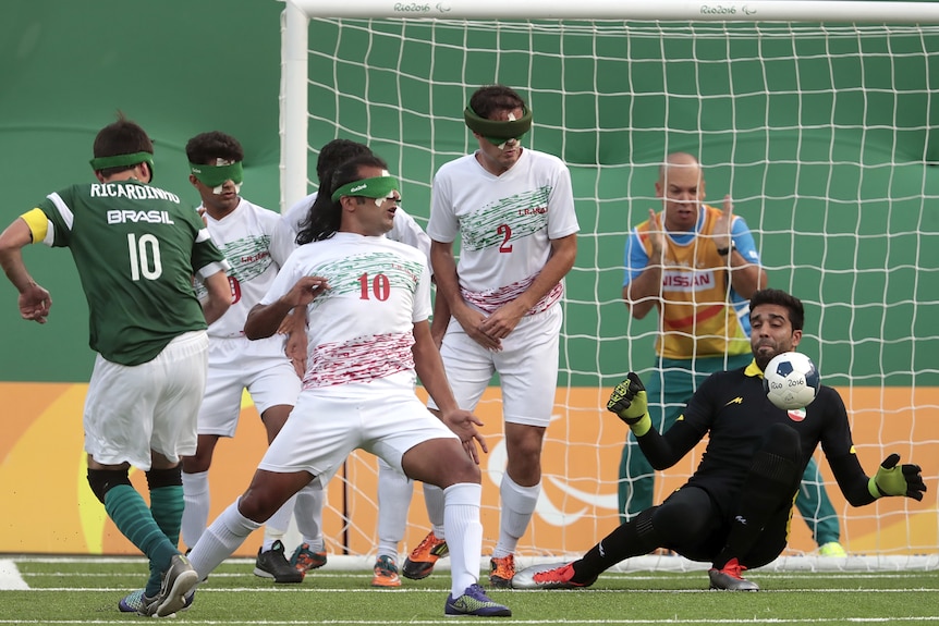 A Brazilian Paralympic footballer wearing blackout goggles takes a shot and the ball goes toward the sighted Iran goalkeeper. 
