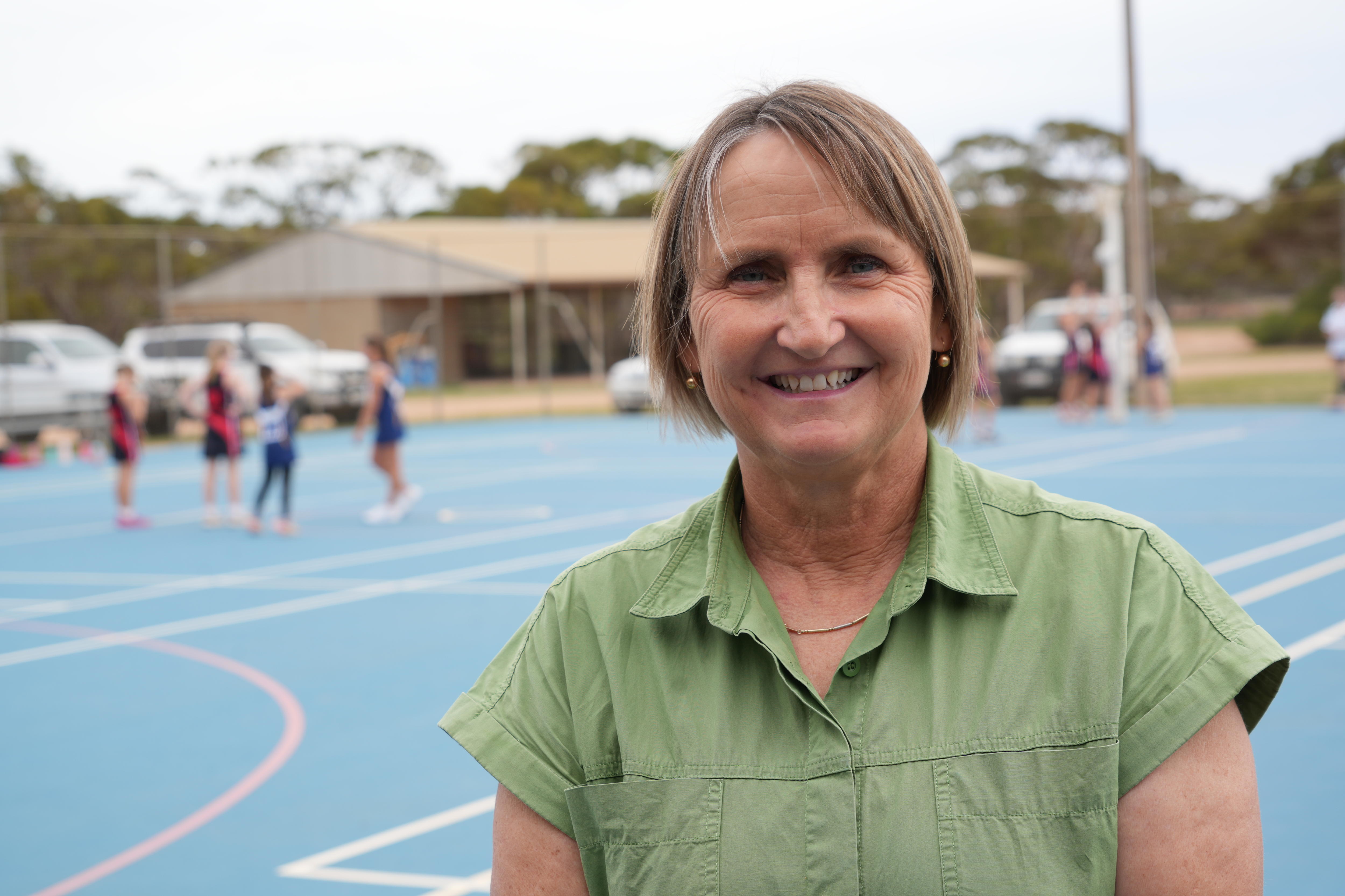 Mulher com top verde em pé na quadra de netball azul com jogadores fora de foco ao fundo