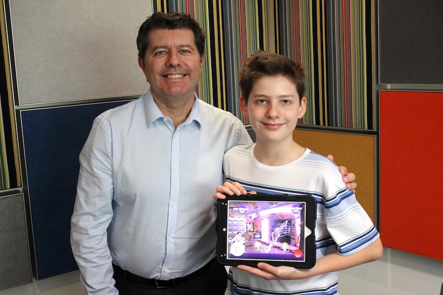 12 year old Hamish Finlayson and hid Dad, Graeme Finlayson, stand in front of a colourful wall holding up an iPad.