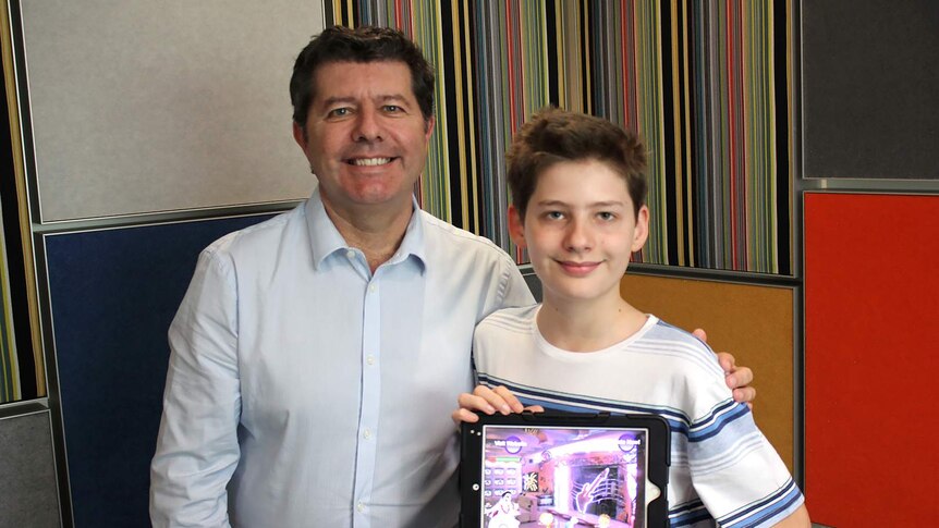 12 year old Hamish Finlayson and hid Dad, Graeme Finlayson, stand in front of a colourful wall holding up an iPad.