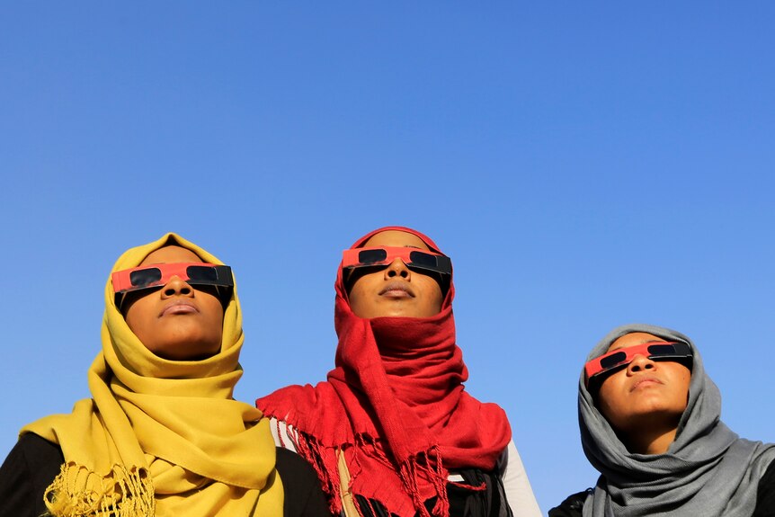 Three women wearing solar glasses looking up at a cloudless blue sky. Each wear headscarves, one yellow, one red, one grey