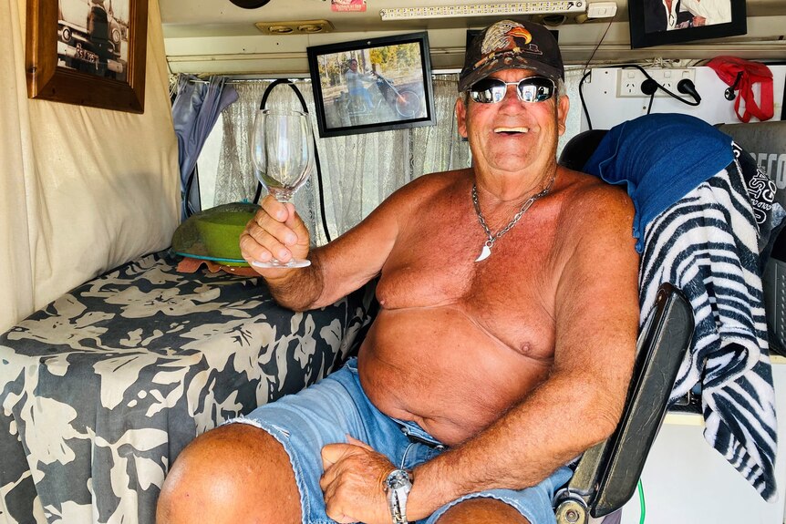Shirtless man sitting inside a mini bus holding a wine glass and smiling.