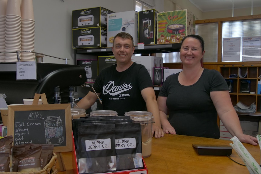A man in a black shirt and a woman in a dark shirt stand behind the counter of a cafe beside a coffee machine.