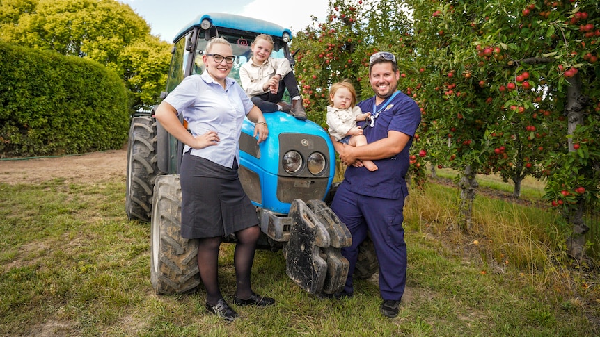 Two adults in nurses uniform lean against a tractor amid apple trees with two kids.
