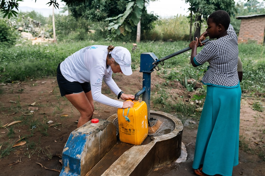 A woman in running gear fills up a water container while another woman in a blue skirt and checked top pumps the well. 