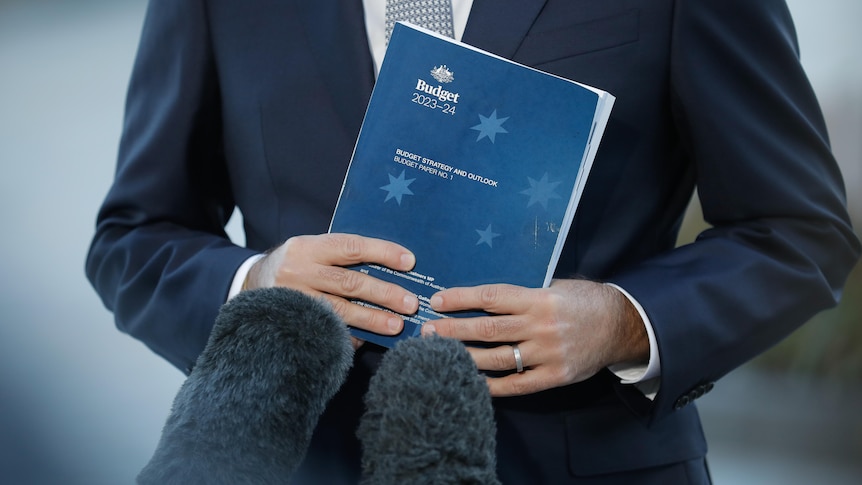 Jim Chalmers holds a thick book with a dark blue cover which a lighter blue Southern Cross titled Budget 2023-24