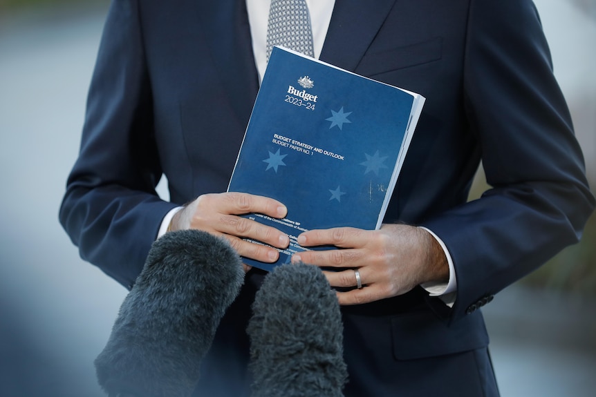 Jim Chalmers holds a thick book with a dark blue cover which a lighter blue Southern Cross titled Budget 2023-24