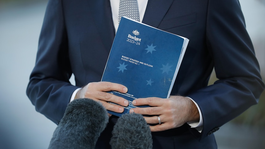 A man wearing a navy suit grasps a blue booklet in two hands, in front of two microphones. The book is titled Budget Paper No. 1