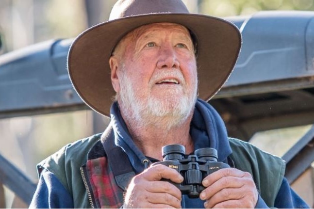 An older man with an Akubra hat looking at the sky holding binoculars