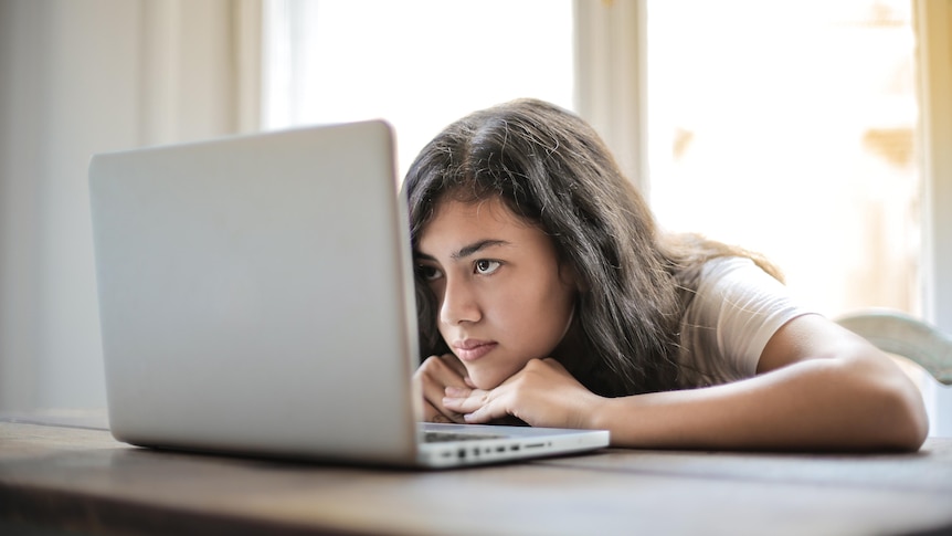 A young woman with dark hair rests her chin on her hands, arms flat on the desk, while staring at her laptop screen