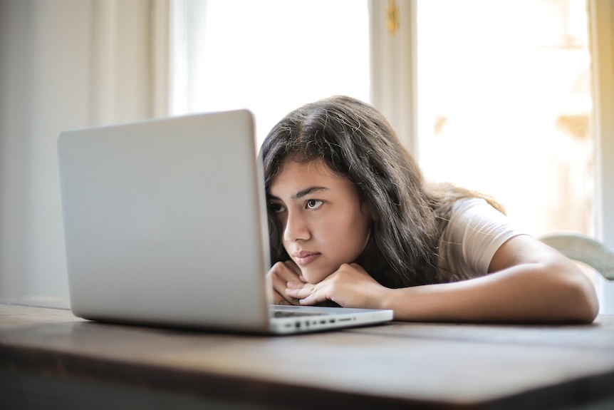 A young woman with dark hair rests her chin on her hands, arms flat on the desk, while staring at her laptop screen