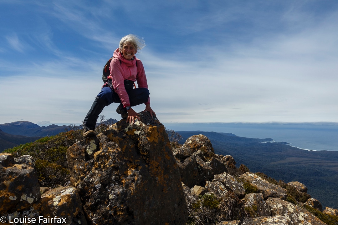 a woman in a pink top on top of a very tall mountain, with ocean and hills in the background