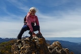 a woman in a pink top on top of a very tall mountain, with ocean and hills in the background