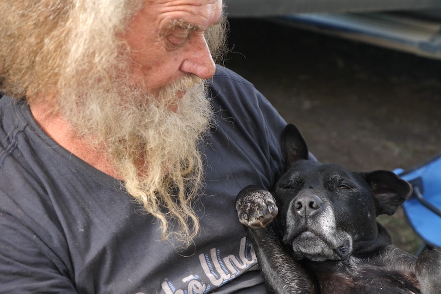 A wild white hair and bearded man looks down at his dog sleeping in his lap, belly up.