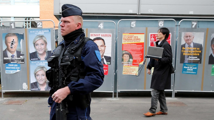 A police officer with gun, a person walks behind him, posters of the candidates in the background.